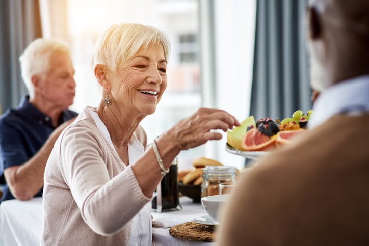 Older woman at a dining table picking a piece of fruit from a serving platter.